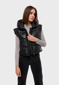 Gilet trapuntato nero con cappuccio, dotato di chiusura con zip e motivo trapuntato. Indossato sopra una maglietta a maniche lunghe grigia chiara e pantaloni neri.