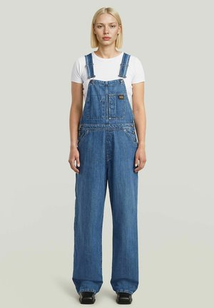 Młoda kobieta o blond włosach, ubrana w biały t-shirt i luźne niebieskie ogrodniczki jeansowe, stojąca na tle jednolitego jasnego tła.