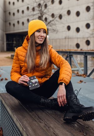 Orange puffer jacket, black skinny jeans, and black lace-up combat boots. Holding a black wallet with polka dot accents, seated on a wooden surface.