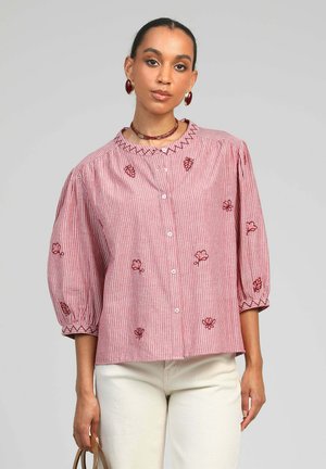 Femme portant un chemisier rose rayé avec des fleurs brodées, des manches trois-quarts bouffantes, et un pantalon blanc, tenant un petit sac marron.