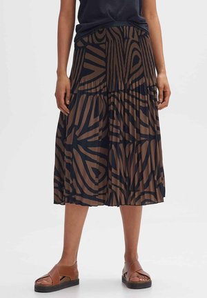 Mujer vistiendo una falda midi plisada de patrón abstracto en marrón y negro, una blusa negra y sandalias de plataforma marrones, de pie sobre un fondo blanco.
