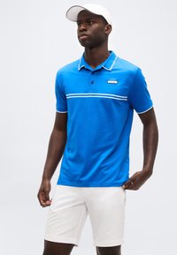 Άνδρας που φοράει μπλε πόλο με λευκές ρίγες και άσπρο καπέλο, συνδυασμένο με άσπρο σορτς, με φόντο ένα απλό τοπίο.