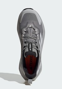 Zapatilla deportiva gris con parte superior de malla texturizada, cierre de cordones, acentos negros y suela de goma. El interior cuenta con una plantilla roja etiquetada como "TERREX."