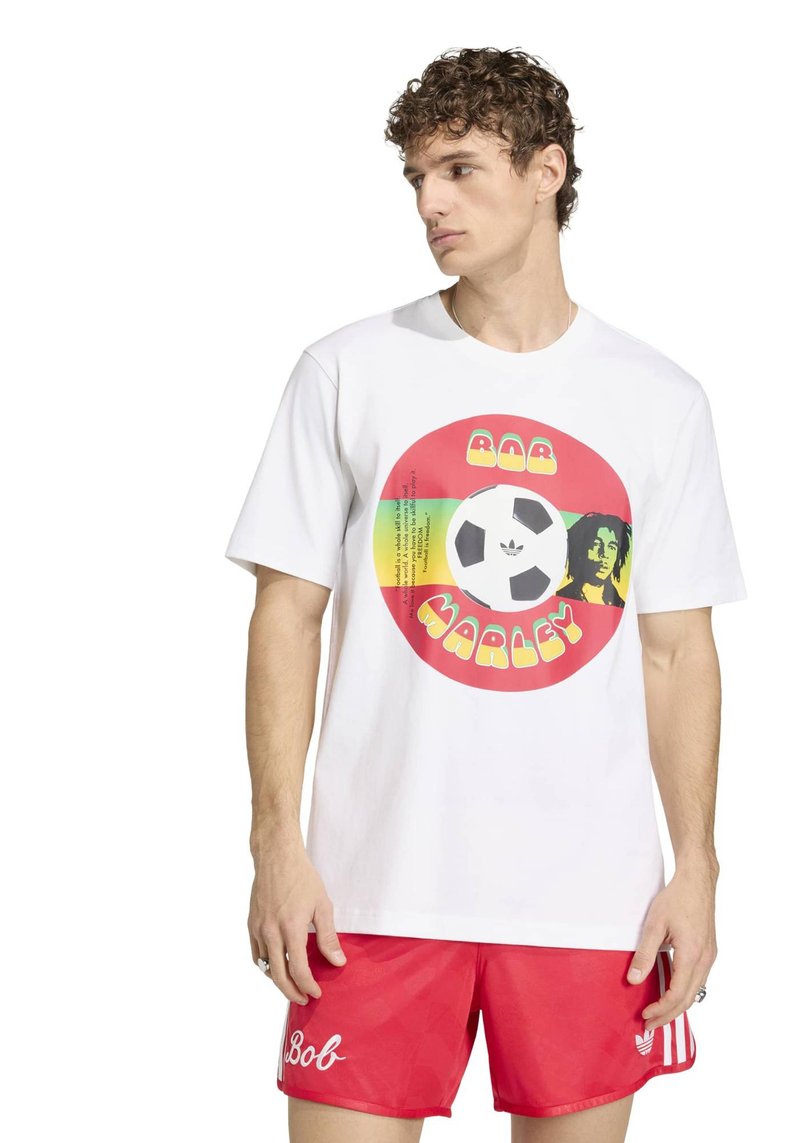Junger Mann mit weißem T-Shirt mit Bob-Marley-Fußballgrafik und roter Shorts mit "Bob"-Text auf dem linken Bein.