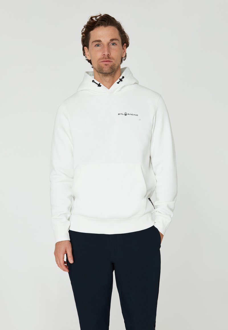 Sudadera blanca con capucha y bolsillo frontal, con un logo negro en el pecho. Textura suave y puños y dobladillo acanalados. Combinada con pantalones oscuros.