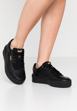 Person, die schwarze Puma-Plateau-Sneaker mit goldenen Logos trägt, steht auf einem glänzenden hellgrauen Boden vor einem schlichten weißen Hintergrund.