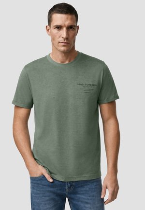 Mężczyzna w zielonym t-shircie z krótkim rękawem z napisem na piersi i niebieskich dżinsach, stojący z jedną ręką w kieszeni na jednolitym tle.