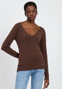 Brauner V-Ausschnitt-Pullover mit langen Ärmeln, geripptem Saum und Bündchen, aus weichem Material gefertigt. Kombiniert mit hellblauen Jeans. Minimales Branding-Detail.