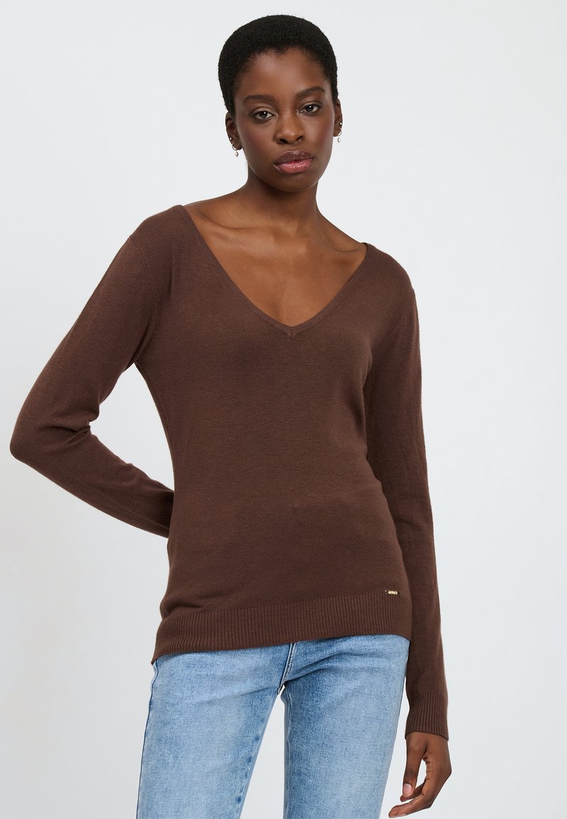 Brauner V-Ausschnitt-Pullover mit langen Ärmeln, geripptem Saum und Bündchen, aus weichem Material gefertigt. Kombiniert mit hellblauen Jeans. Minimales Branding-Detail.