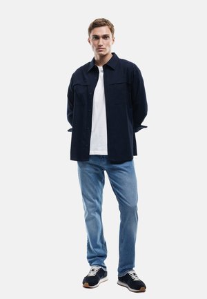 Donkerblauwe overhemd met knopen, witte t-shirt eronder, lichtblauwe jeans, en marineblauwe sneakers met witte veters en bruine zolen, staande op een effen achtergrond.