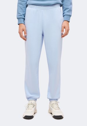 Personne portant un pantalon de jogging bleu clair avec des poignets élastiques et des baskets blanches, debout devant un fond uni.