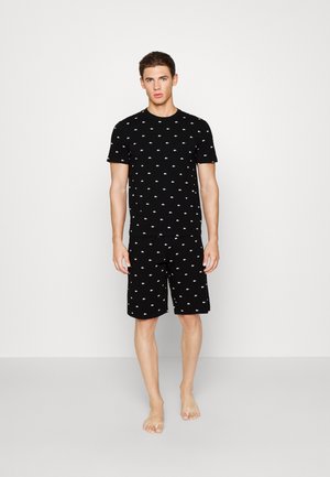 Joven de pie descalzo, con una camisa negra de manga corta y pantalones cortos a juego con pequeños patrones de logotipos blancos por todas partes.