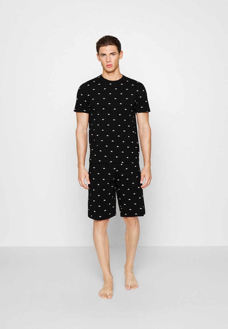 Junger Mann steht barfuß und trägt ein schwarzes kurzärmeliges Hemd sowie passende Shorts mit kleinen weißen Logomustern überall.