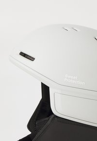Biely prilba s ventilačnými otvormi a čiernymi akcentmi, na boku s nápisom "Sweet Protection", pripevnená na čiernom podklade.