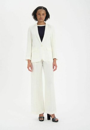 Blazer sur mesure de couleur claire et pantalon large en tissu texturé. Des chaussures à plateforme noires complètent l'ensemble.
