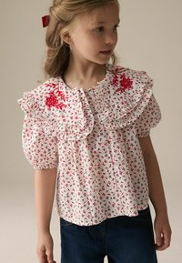 Weiße Bluse mit floralem Muster und Rüschensaum, mit roter Stickerei und kleinen Blumenmustern. Kurze Puffärmel und Knopfleiste.