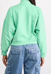 Felpa verde menta con collo alto e spalle scese, abbinata a pantaloni cargo azzurri chiari con tasche laterali e posteriori.