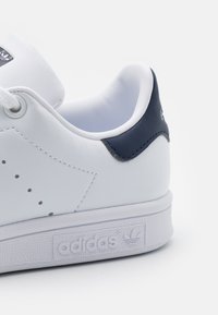 Vit lädersneaker med marinblå hälflik, perforerat mönster på sidorna och texturerad vit gummisula med Adidas-logotypen.