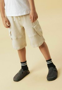 Shorts cargo beiges avec deux poches latérales, associés à des chaussettes noires à rayures blanches et des chaussures à enfiler grises. Design simple et décontracté.