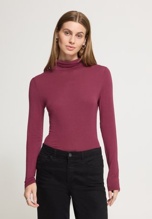 Femme aux cheveux lisses châtain clair porte un col roulé prune ajusté à manches longues et un pantalon noir, debout devant un fond uni clair.