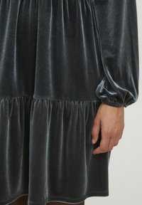 Grå sammetsklänning med en upptagen kjol, långa ärmar och elastiska manschetter. Mjuk textur med en flödande silhuett och lagerdesign.