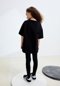 Črna prevelika majica, črne legice z belim tiskom na strani in črno-bele superge. Od strani je videti kodraste lase. Minimalistično ozadje.