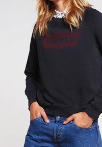 Black sweatshirt with red Levi's logo, crew neckline, and long sleeves. Paired with blue jeans, fabric appears soft and textured.