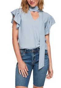 Femme portant un chemisier bleu clair à manches volantées avec un nœud au cou et un short en denim bleu longueur genou, posant les mains détendues.