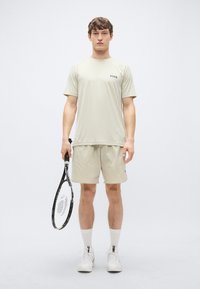 Αθλητική μπλούζα ανοιχτού γκρι χρώματος με κοντά μανίκια και μικρό λογότυπο. Συνδυάζεται με αντίστοιχα σορτς και λευκά παπούτσια τένις, κρατώντας μια μαύρη ρακέτα τένις.