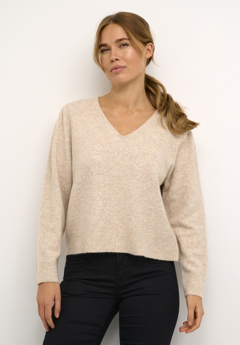Bež pulover z v-neck iz soft materiala z teksturo. Ima dolge rokave in sproščen kroj, v kombinaciji s temnimi oprijetimi hlačami.