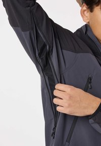 Persona parzialmente visibile che slaccia la cerniera ventilata laterale di una giacca grigio scuro e nera, mostrando il pannello in rete sottostante.