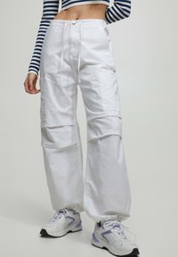 Vrouw in witte cargo broeken met een trekkoord in de taille en witte sneakers met paarse accenten, gecombineerd met een navy en witte gestreepte lange mouw crop top.