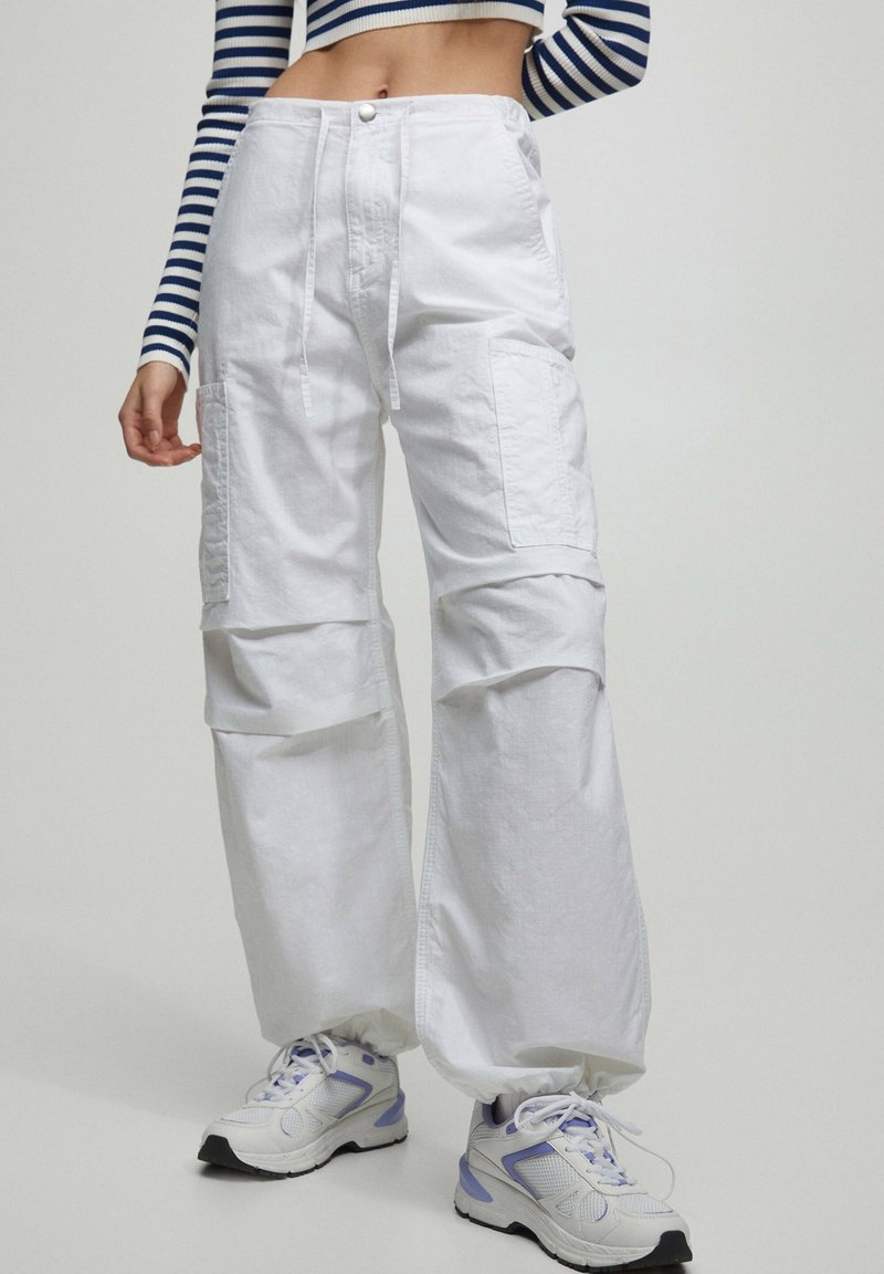 Vrouw in witte cargo broeken met een trekkoord in de taille en witte sneakers met paarse accenten, gecombineerd met een navy en witte gestreepte lange mouw crop top.