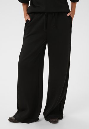 Personne portant un pantalon noir ample avec poches latérales et chaussures foncées, debout les mains dans les poches devant un fond uni.