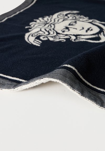 Σκούρα μπλε πετσέτα με λευκό σχέδιο με κεφάλι Μέδουσας και λευκό περιθώριο με ρίγες, μερικώς διπλωμένη πάνω σε ανοιχτόχρωμη επιφάνεια.