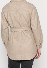Cappotto di lana beige con cintura e maniche lunghe indossato su pantaloni neri, mostrato di spalle su uno sfondo chiaro e semplice.