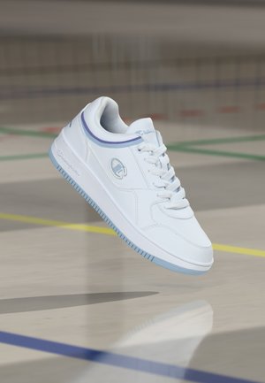 Sneaker Champion bianco con suola azzurro chiaro e dettagli viola sospesa sopra un pavimento da palestra segnato con linee colorate.