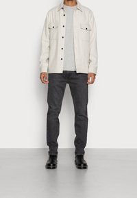 Hombre vistiendo una chaqueta de botón color beige claro sobre una camisa gris, jeans gris oscuro y botas de cuero negras, de pie contra un fondo blanco.