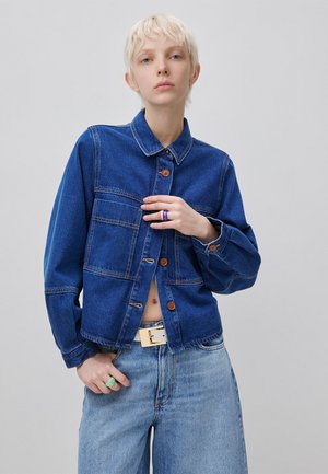 Persona con capelli corti e biondi che indossa una giacca di jeans blu scuro abbottonata, jeans azzurri e anelli su entrambe le mani su uno sfondo semplice.