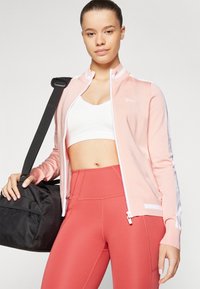 Világos rózsaszín zipzáras dzseki fehér részletekkel, bordázott textúrával és logóval. Egy fehér crop top felett viseli, rózsaszín, magas derekú leggings-szel, miközben egy fekete táskát tart.
