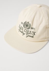 Gorra de béisbol en tela crema con visera curvada. Presenta texto bordado en verde y un emblema circular, con detalles de costura visibles.