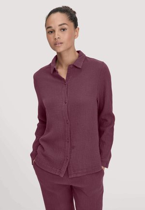 Giovane donna con le mani nelle tasche, indossando una camicia a maniche lunghe bordeaux con bottoni e pantaloni abbinati, su uno sfondo neutro.