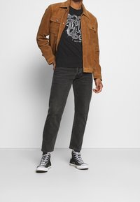 Chaqueta de ante marrón sobre una camiseta gráfica negra, combinada con jeans oscuros y zapatillas altas negras. Diseño simple y colores contrastantes.