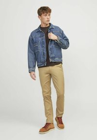 Denim takki keski sinisessä, ruskea pyöreä kaula-aukko paita, khakihousut ja vaaleat saappaat punaisilla nauhoilla; seisova asento käsi takin päällä.