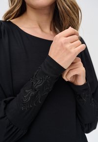 Kobieta w czarnej bluzce z długim rękawem, z misternymi wytłaczanymi wzorami kwiatowymi na mankietach, poprawiająca jeden rękaw przy ramieniu.