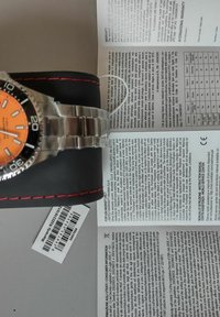 Zilveren metalen horloge met een oranje wijzerplaat en zwarte bezel, geplaatst op een zwart kussen met rode stiksels. Garantiepapier is gedeeltelijk zichtbaar.
