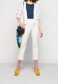 Kvinna som bär vita cropped byxor, gula klackar, marinblå topp, vit kofta, håller en svart handväska med en färgglad scarf mot en enfärgad bakgrund.