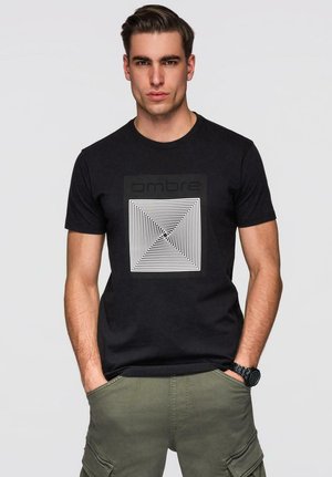 Homme aux cheveux lissés en arrière portant un t-shirt noir avec un motif graphique et un pantalon cargo vert, les mains dans les poches, sur un fond gris uni.
