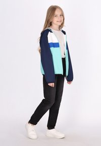 Farverig vindjakke med marineblå, turkise og hvide paneler, der har en lynlåslukning og hætte, parret med sorte jeans og hvide sneakers.