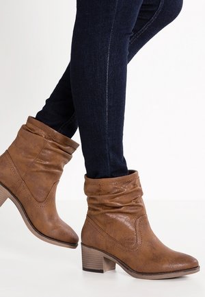 Žena v tmavě modrých úzkých džínách a hnědých volných kotníkových botách na vysokém robustním podpatku na bílém pozadí.